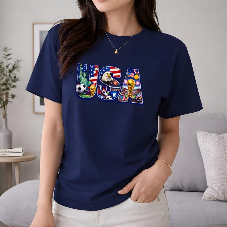 May include: Navy blue t-shirt with a colorful "USA" graphic. The graphic incorporates images of the Statue of Liberty, an American flag, a bald eagle, soccer balls, and a soccer trophy. The shirt is a classic crew neck style.