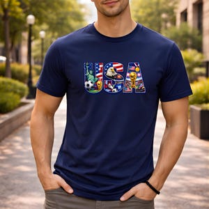 May include: Navy blue t-shirt with the word "USA" in a colorful design featuring the Statue of Liberty, an eagle, a soccer ball, and a trophy. The shirt is made of a soft material.