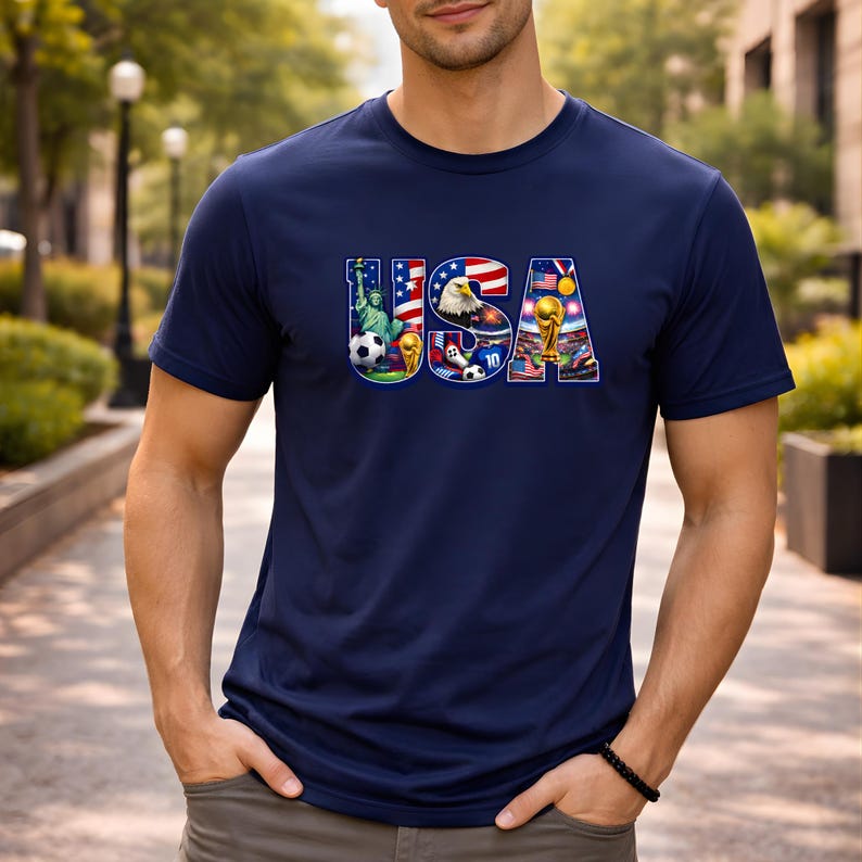 May include: Navy blue t-shirt with the word "USA" in a colorful design featuring the Statue of Liberty, an eagle, a soccer ball, and a trophy. The shirt is made of a soft material.