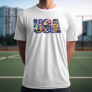 May include: White t-shirt with a colorful graphic of the word "USA" filled with images of the Statue of Liberty, an eagle, soccer balls, and a stadium with a trophy. The shirt is made of a lightweight material.