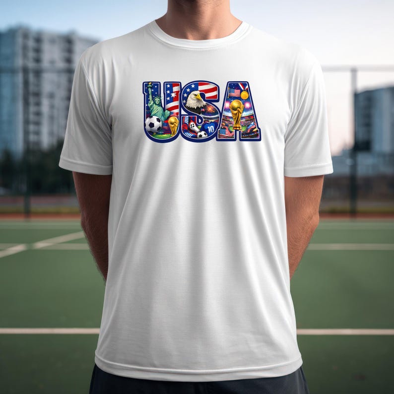 May include: White t-shirt with a colorful graphic of the word "USA" filled with images of the Statue of Liberty, an eagle, soccer balls, and a stadium with a trophy. The shirt is made of a lightweight material.