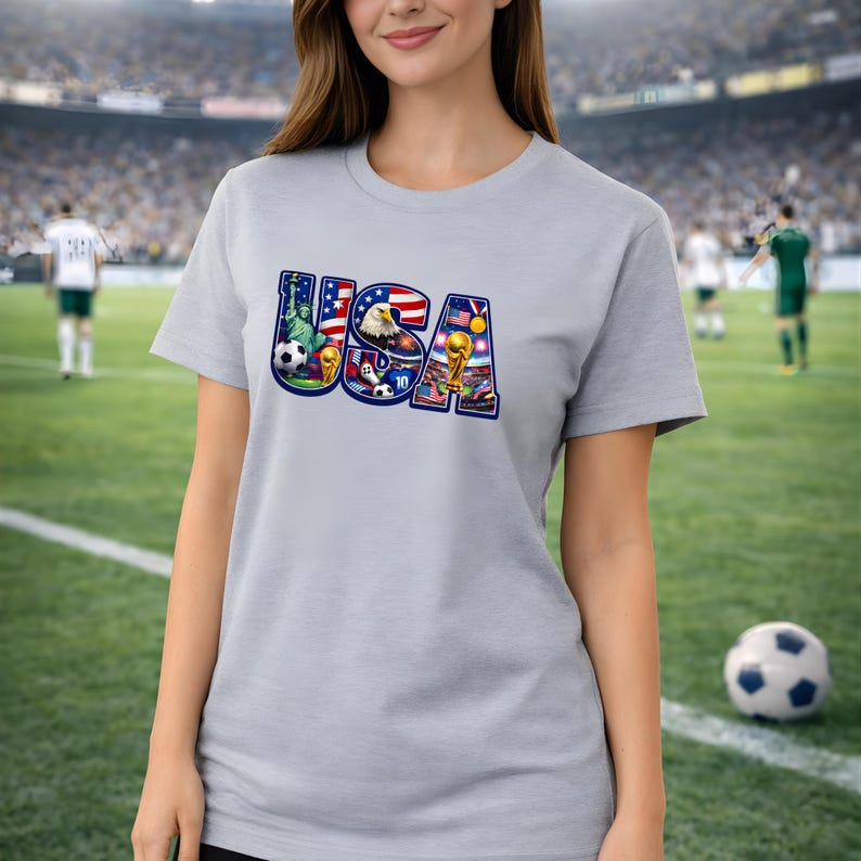 May include: A light gray t-shirt featuring the word "USA" in a colorful design. The design incorporates elements like the Statue of Liberty, an eagle, soccer balls, and a trophy, all set against the backdrop of a soccer field.