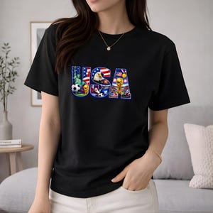 May include: Black t-shirt featuring the word "USA" in a colorful design. The design incorporates elements like the Statue of Liberty, an eagle, soccer balls, and a trophy, all set against the backdrop of the American flag.