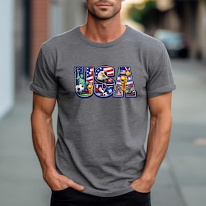 May include: Gray t-shirt with the word "USA" in a colorful design. The design incorporates elements like the Statue of Liberty, an eagle, soccer balls, and a trophy, all set against the backdrop of the American flag.