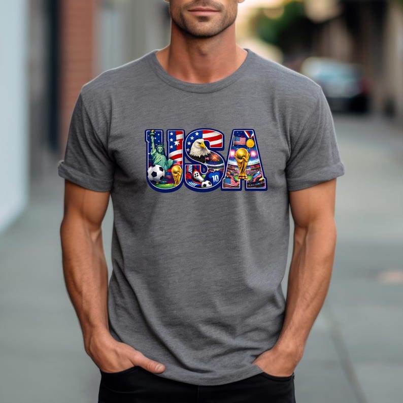 May include: Gray t-shirt with the word "USA" in a colorful design. The design incorporates elements like the Statue of Liberty, an eagle, soccer balls, and a trophy, all set against the backdrop of the American flag.