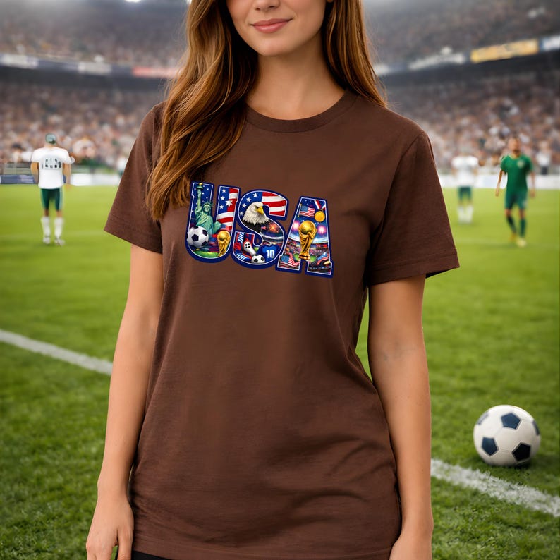 May include: A brown t-shirt featuring the word "USA" in a colorful design. The design incorporates elements like the American flag, an eagle, soccer balls, and the Statue of Liberty. The shirt is worn by a person in a soccer stadium.