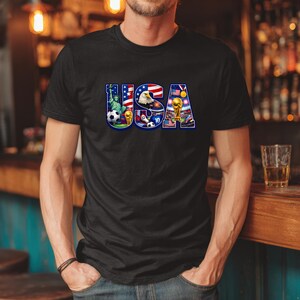 May include: Black t-shirt with a colorful "USA" graphic. The graphic incorporates elements like the Statue of Liberty, an eagle, a soccer ball, and a trophy, all in patriotic colors. The shirt is a casual wear item.