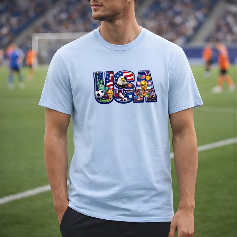 May include: Light blue t-shirt featuring the word "USA" in a colorful design. The design incorporates elements like a soccer ball, the Statue of Liberty, an eagle, and a soccer trophy, all in vibrant colors.