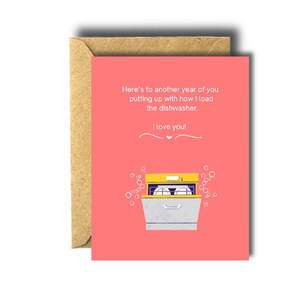 May include: A greeting card with a coral background and a cartoon illustration of a dishwasher. The card reads, "Here's to another year of you putting up with how I load the dishwasher. I love you!" The card is inside a brown envelope.