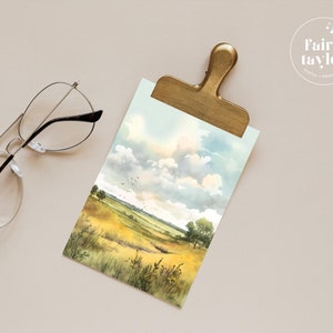 May include: A watercolour painting of a field with tall grass and a cloudy sky. The painting is on a white background and is framed by a gold clip.