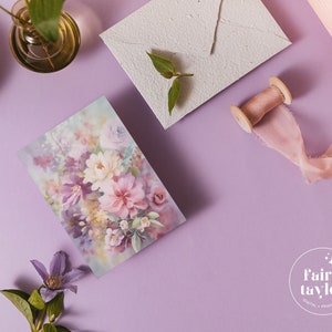May include: A floral print card featuring a watercolor design of pink, purple, and white flowers. The card is set on a lavender surface with a matching envelope, a spool of pink ribbon, and a small glass vase with greenery.