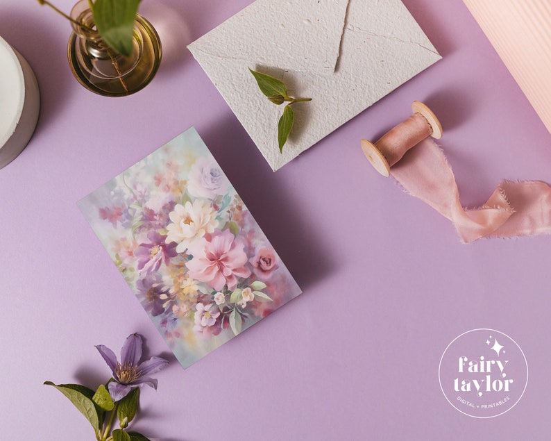 May include: A floral print card featuring a watercolor design of pink, purple, and white flowers. The card is set on a lavender surface with a matching envelope, a spool of pink ribbon, and a small glass vase with greenery.