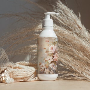 May include: White pump bottle with a floral design featuring pink roses and white blossoms. The bottle has a white pump dispenser and a decorative label with a vintage-style floral pattern. The background includes dried ornamental grass.
