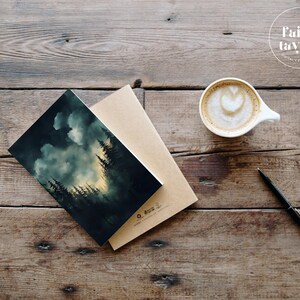 May include: A dark green and blue watercolour illustration of a forest with a cloudy sky. The illustration is printed on a card and is sitting on a brown envelope. The card is on a wooden table with a cup of coffee and a pen.