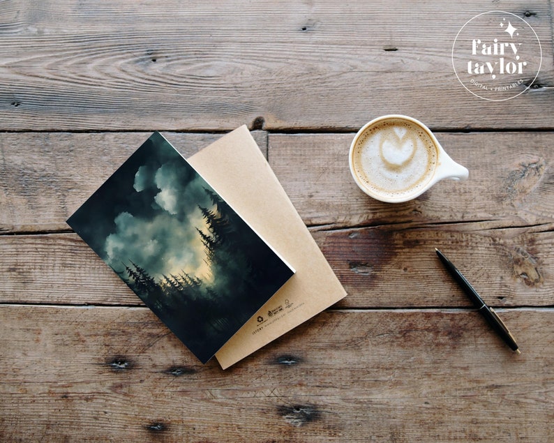 May include: A dark green and blue watercolour illustration of a forest with a cloudy sky. The illustration is printed on a card and is sitting on a brown envelope. The card is on a wooden table with a cup of coffee and a pen.