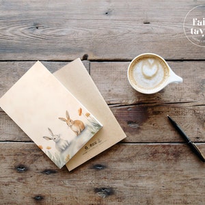 May include: A brown and white card with a watercolor illustration of two rabbits in a field of orange flowers. The card is sitting on a brown envelope and a wooden table. There is a cup of coffee and a pen on the table.