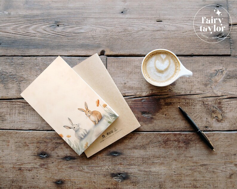May include: A brown and white card with a watercolor illustration of two rabbits in a field of orange flowers. The card is sitting on a brown envelope and a wooden table. There is a cup of coffee and a pen on the table.