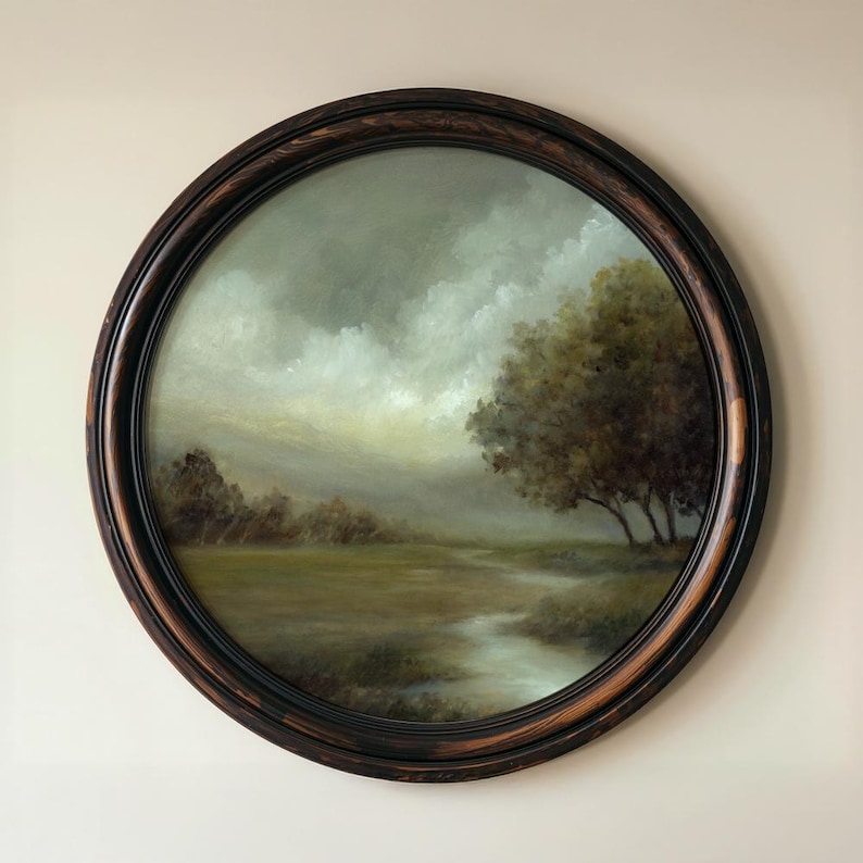 May include: A round oil painting in a dark wood frame depicting a landscape with a grassy field, a winding stream, and a cluster of trees under a cloudy, overcast sky. The artwork features muted green, brown, and grey tones with soft lighting.