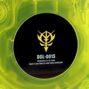 Może przedstawiać: Żółta konsola GameCube z czarną osłoną napędu dysków. Osłona zawiera złoty emblemat ze stylizowanym wzorem i tekstem "DOL-001S KSIĘSTWO ZEON SPERSONALIZOWANA KONSOLE NINTENDO GAMECUBE CHARA".