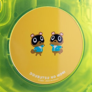 May include: A yellow disc featuring two cartoon tanuki characters with black eyes and brown ears, wearing blue jackets. The disc has the text "DOUBUTSU NO MORI" and copyright information. The disc is set inside a transparent green case.