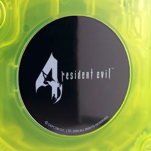 Op de afbeelding: Een zwarte schijf met een zilveren "4" en de tekst "Resident Evil" in wit. De schijf is omgeven door een felgele plastic behuizing.