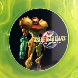 Puede incluir: Un disco de juego de GameCube amarillo con un círculo negro en el centro. El círculo presenta un personaje de videojuego con un traje verde y naranja, sosteniendo un arma. El texto "METROID PRIME" está impreso en blanco en el círculo.