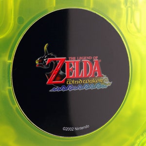 May include: A close-up of a green GameCube game disc with a black label. The label features the logo for "The Legend of Zelda: The Wind Waker" with the text "©2002 Nintendo".