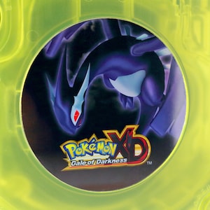Könnte beinhalten: Ein gelbes Game Boy Advance SP-Spielecase mit einer schwarzen und violetten Pokemon XD Gale of Darkness-Spielkassette im Inneren.