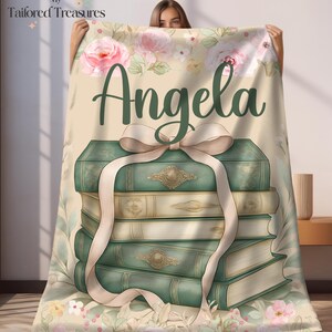 Op de afbeelding: Een gepersonaliseerde deken met een stapel boeken in vintage-stijl, vastgebonden met een cr&egrave;mekleurig lint. De naam "Angela" staat in donkergroen geschreven. De achtergrond heeft een bloemenontwerp met roze rozen en groene bladeren.