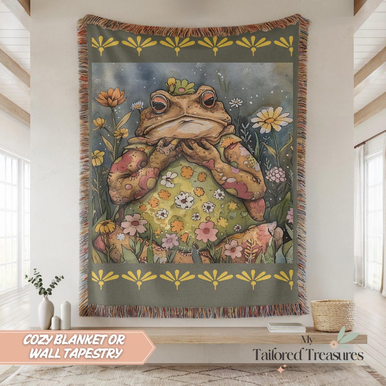May include: A woven tapestry or blanket featuring a whimsical illustration of a frog in a floral dress, surrounded by flowers. The artwork has a vintage aesthetic with a muted color palette of greens, yellows, and pinks. The text "COZY BLANKET OR WALL TAPESTRY" is visible.
