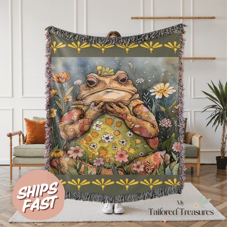 May include: A woven throw blanket featuring a whimsical illustration of a frog in a floral meadow. The frog is adorned with flowers and surrounded by colorful blooms. The blanket has a green border with yellow floral accents and fringed edges. The words "SHIPS FAST" are visible.