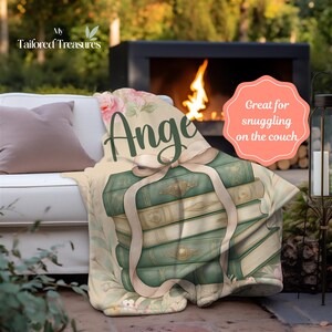 Op de afbeelding: Een beige deken met een ontwerp van groene boeken die met een beige lint zijn vastgebonden. De deken heeft een bloemen- en botanisch ontwerp met de naam "Ange" in groen schrift. De deken ligt op een witte bank met een kussen.