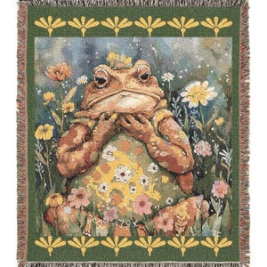 May include: A woven throw blanket featuring a whimsical illustration of a frog in a floral setting. The frog is brown and wears a crown, surrounded by colorful flowers. The border is green with a repeating yellow floral pattern.