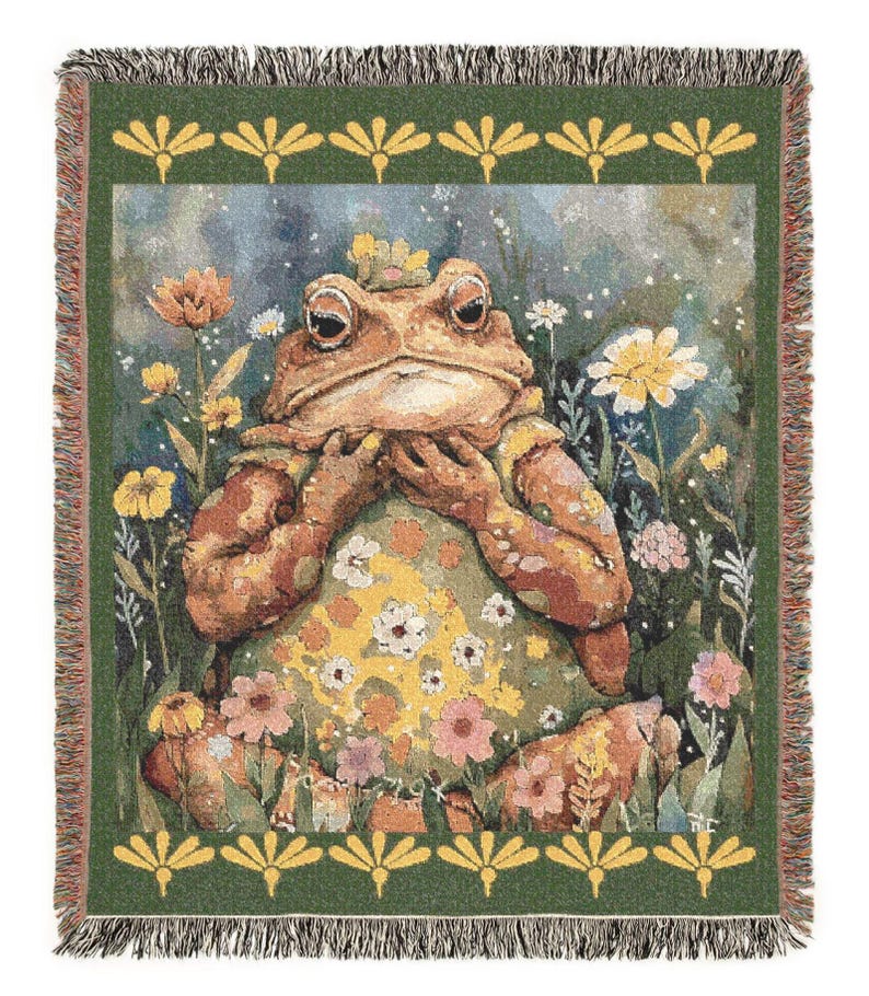 May include: A woven throw blanket featuring a whimsical illustration of a frog in a floral setting. The frog is brown and wears a crown, surrounded by colorful flowers. The border is green with a repeating yellow floral pattern.