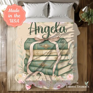 Op de afbeelding: Een gepersonaliseerde deken met een stapel boeken in vintage-stijl, vastgebonden met een lichtroze lint. De naam "Angela" staat in groene letters boven de boeken. De deken heeft een bloemen- en botanisch ontwerp.