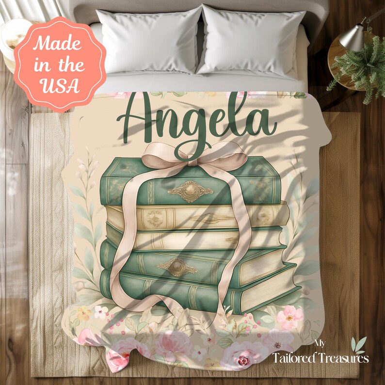 Op de afbeelding: Een gepersonaliseerde deken met een stapel boeken in vintage-stijl, vastgebonden met een lichtroze lint. De naam "Angela" staat in groene letters boven de boeken. De deken heeft een bloemen- en botanisch ontwerp.