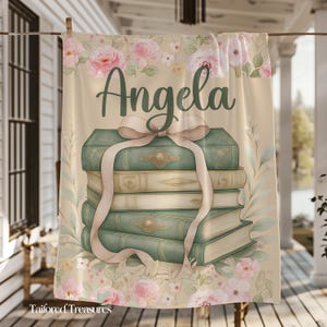 Op de afbeelding: Een beige handdoek met een aquarelontwerp met een stapel boeken vastgebonden met een roze lint. Het woord "Angela" staat in script boven de boeken, omringd door roze rozen en groene bladeren.