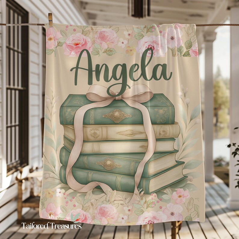 Op de afbeelding: Een beige handdoek met een aquarelontwerp met een stapel boeken vastgebonden met een roze lint. Het woord "Angela" staat in script boven de boeken, omringd door roze rozen en groene bladeren.