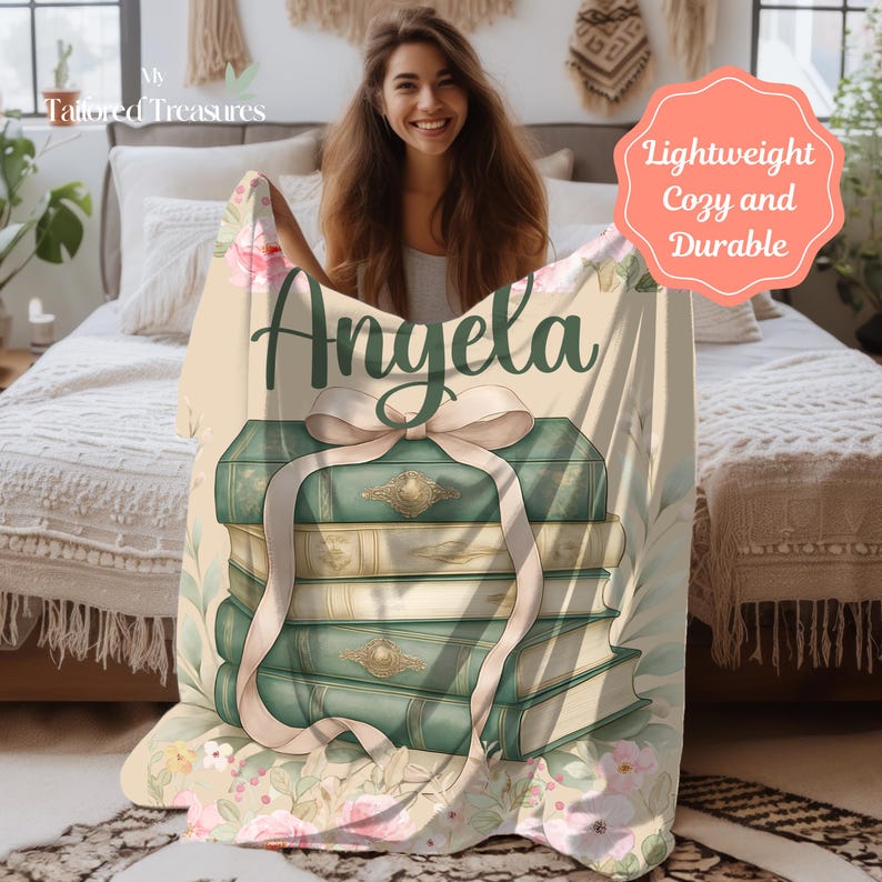 Op de afbeelding: Een beige deken met een ontwerp van groene en gouden boeken die met een roze lint zijn vastgebonden. De naam "Angela" staat in groene letters boven de boeken. De deken heeft een bloemenpatroon en de woorden "Lightweight, Cozy and Durable".