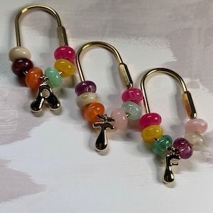 May include: Three gold-toned U-shaped metal clasps, each adorned with a variety of colorful, smooth, round beads in shades of pink, yellow, orange, green, and purple. Each clasp features a dangling gold-toned letter charm: A, T, and F.