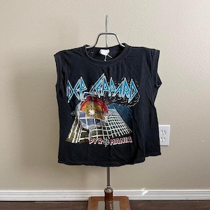 Könnte beinhalten: Schwarzes ärmelloses T-Shirt mit einem Grafikdesign mit dem Bandnamen Def Leppard in Blau und dem Wort "Pyromania". Die Grafik enthält ein Pyramiden-Design mit einem farbenfrohen, kreisförmigen Element.