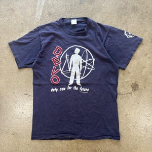 May include: A navy blue short-sleeve t-shirt featuring a white graphic of a figure inside a circular atom design. The word DEVO is printed in red vertically, with the text duty now for the future in white below. A small white graphic is on the sleeve.