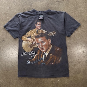 May include: A charcoal gray short-sleeve t-shirt featuring a graphic print of Elvis Presley in a military uniform. The design includes a portrait of Elvis, a depiction of him playing an acoustic guitar, and the white printed signature Elvis Presley.