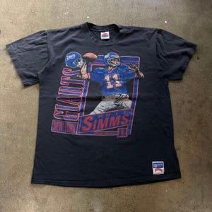 May include: A black t-shirt featuring a graphic of a football player in a blue uniform throwing a football. The shirt has the words "Giants" and "Simms" in red and blue, with "New York" below. A small tag is on the bottom right.