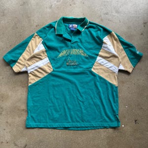 May include: Teal and tan polo shirt with short sleeves. The shirt features a white and tan color block design on the sleeves and sides. The text "GOLD MINERS" is embroidered on the chest in gold thread.