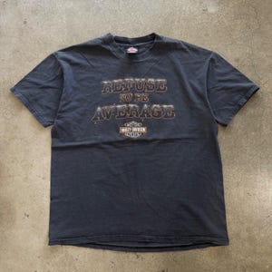 Może przedstawiać: Czarny t-shirt z krótkim rękawem z napisem "REFUSE TO BE AVERAGE" wykonanym stylizowaną czcionką. Poniżej tekstu znajduje się pomarańczowo-białe logo Harley-Davidson Motor Cycles. Koszulka ma okrągły dekolt i standardowy krój.