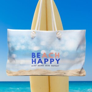 May include: A white beach tote bag with a blue and pink graphic that says "Beach Happy Surf Sand Sun Repeat" on a surfboard against a blue sky and ocean background.