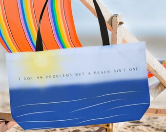 Könnte beinhalten: Eine blaue und weiße Einkaufstasche mit dem Text "I got 99 problems but a beach ain't one" auf der Vorderseite. Die Tasche liegt auf einem Strandstuhl mit einem regenbogenfarbenen Stoff.