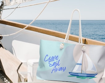 May include: A white canvas tote bag with blue text that reads "Come Sail Away" and a blue and white sailboat graphic. The bag is on a sailboat with a white sail and a wooden mast.