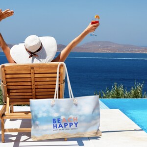 May include: A woman relaxes in a lounge chair by a pool with a blue and white beach bag that says "Beach Happy Surf Sand Sun Repeat".